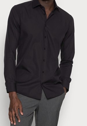 Mann trägt ein schwarzes, langärmeliges Hemd mit Knöpfen und graue Hose, eine Hand in der Tasche, steht vor einem einfarbigen hellen Hintergrund.