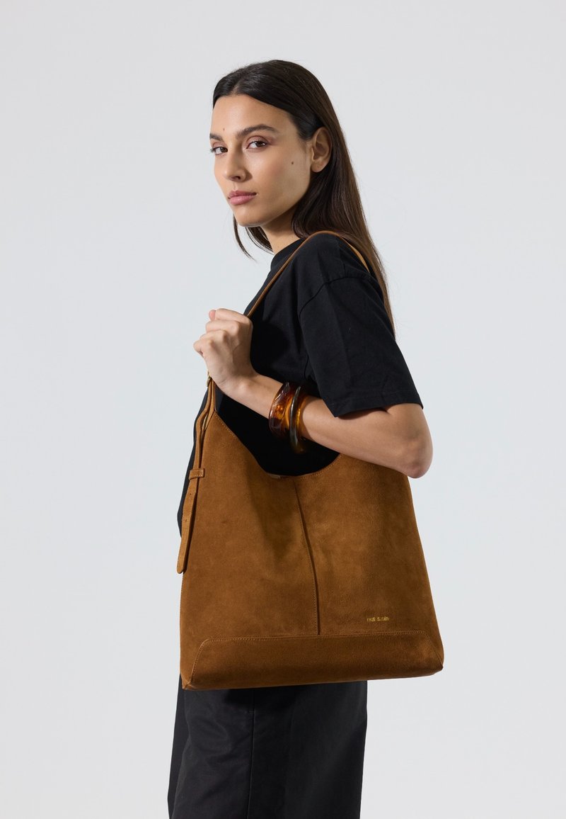Femme aux longs cheveux foncés portant une robe noire et des bracelets en ambre, portant un grand sac à bandoulière en daim marron sur son épaule droite.