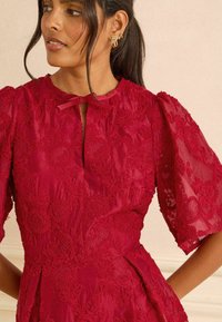 Punase tikandiga kleit puff varrukatega, võtmeaukude kaelus ja paeladetail. Omab lillemustreid ja tekstuurset kangast.