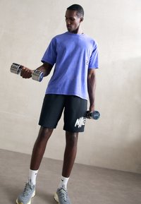 Man in purple shirt and black Nike shorts lifting 5kg and 6kg dumbbells indoors against a plain wall.