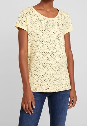 Donna con una maglietta giallo pallido a maniche corte con piccoli motivi a triangolo neri, abbinata a jeans blu, su uno sfondo grigio uniforme.