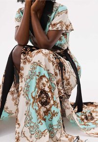 Robe longue en turquoise, noir et crème avec des motifs bruns ornés. Présente un design enroulé et un nœud noir. Tissu doux à motifs avec des manches froufrous.