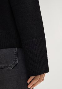 Maglione nero a maglia con dettagli a coste sui polsini e sui polsi. Il materiale sembra morbido con una finitura texture. Jeani grigi visibili nella parte inferiore.