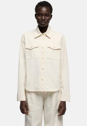 Dunkelhäutige Person trägt cremefarbene Jacke mit Knöpfen und Klappentaschen sowie passende Hose, stehend vor einem schlichten weißen Hintergrund.