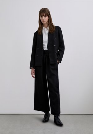 Blazer a righe nere con bottoni doppiopetto, abbinato a pantaloni a gamba larga e una camicia bianca con bottoni. Stivaletti neri completano il look.