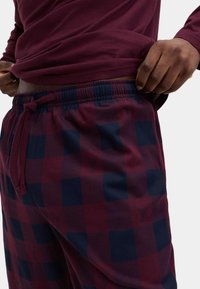 Homme portant une chemise à manches longues bordeaux et un pantalon à carreaux bordeaux et bleu marine, ajustant la ceinture avec la main.