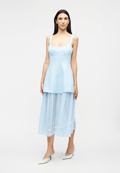 Femeie cu păr lung și întunecat, purtând o rochie bleu deschis fără mâneci, cu o fustă diafană, brodată cu flori, și pantofi albi cu toc.