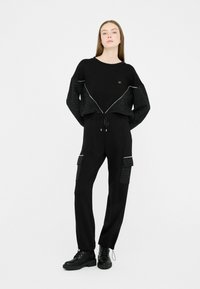 Zwarte sweatshirt met gestructureerde zwarte mouwen, afgewerkt met gouden accenten. Gecombineerd met zwarte broek met cargozakken en een trekkoord in de taille.