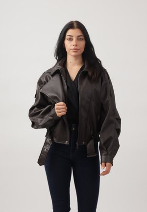 Vrouw met lang donker haar die een donkerbruine oversized leren jas draagt, een zwarte top en donkerblauwe jeans, staand tegen een lichte achtergrond.