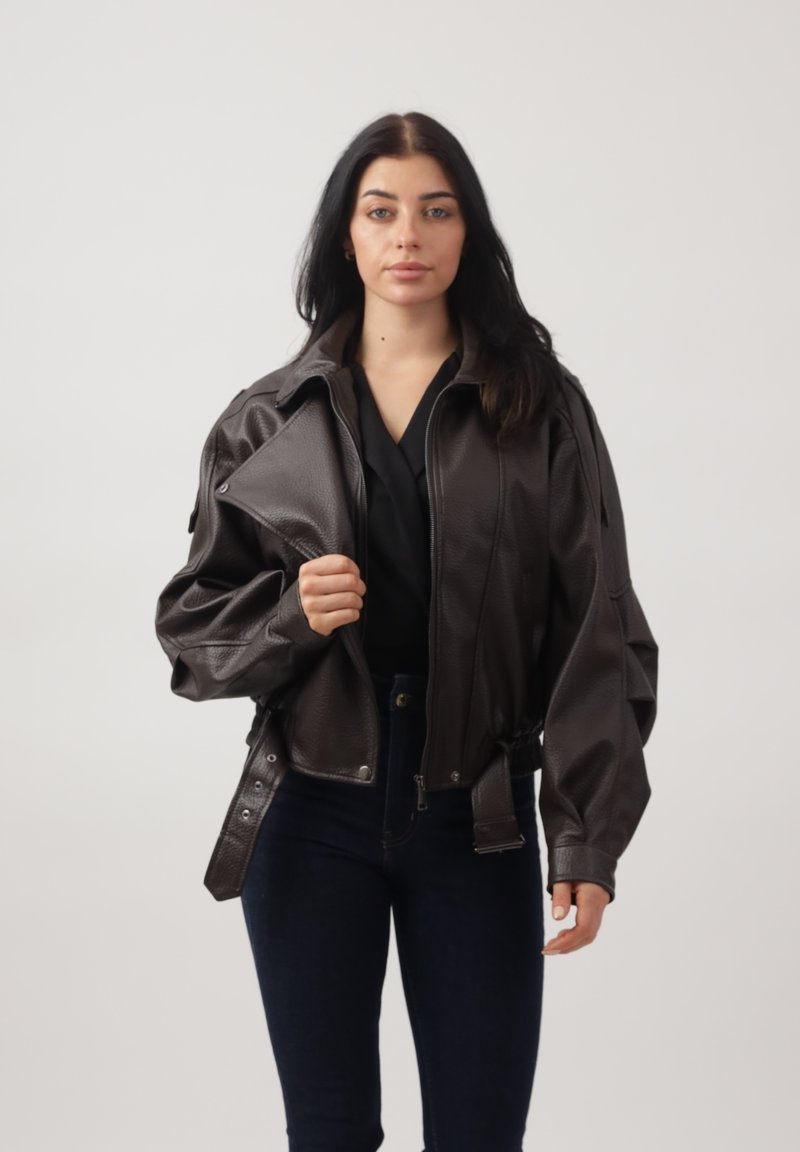 Mujer de cabello largo y oscuro, vistiendo una chaqueta de cuero marrón oscuro de gran tamaño, camiseta negra y vaqueros azul oscuro, de pie frente a un fondo claro.