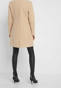 Cappotto beige texture con taglio dritto, maniche lunghe e spacco posteriore, abbinato a legging neri attillati e stivaletti con tacco.