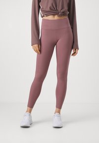 Femme portant un legging taille haute mauve, des baskets blanches et un haut mauve à manches longues noué à la taille, se tenant devant un fond uni.