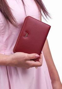 Red leather wallet with gold hardware detail, zip closure, and a smooth texture, being held against a pink fabric backdrop.