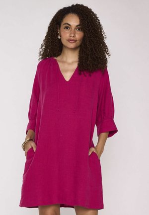 Donna con capelli ricci che indossa un vestito magenta largo, lungo fino al ginocchio, con tasche, maniche a tre quarti e scollo a V.
