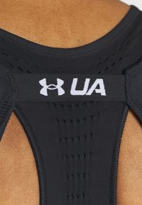 Černé atletické tílko s texturovaným síťovaným vzorem na zádech a bílým logem UA na elastickém pásku. Hladká tkanina a střih.