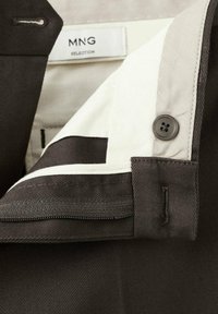 Intérieur d'une veste marron avec doublure crème, équipé d'un bouton et d'une fermeture éclair. L'étiquette indique "MNG SELECTION." Tissu texturé avec des motifs diagonaux.