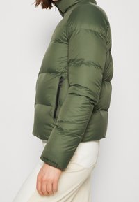 Kurtka puchowa w oliwkowej zieleni z wysokim kołnierzem, boczną kieszenią na suwak, pikowaną teksturą i elastycznymi mankietami. Nazwa marki widoczna na rękawie.