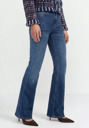 Žena nosí modré bootcut džínsy, tmavé špicaté vysoké opätky a textúrovanú viacfarebnú bundu s dlhými rukávmi s ozdobnými strapcami.