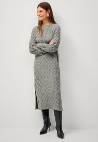 Robe en tricot grise avec un motif en torsades, manches longues, longueur mi-mollet et fente sur le côté. Assortie à des bottes à talons hauts noires.