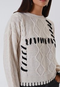 Maglione lavorato a maglia crema con motivo a trecce e dettagli neri, dal taglio rilassato, con scollatura rotonda e orlo a coste. Tessuto con motivi unici.