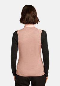 Persona con capelli corti e marroni indossa un gilet senza maniche rosa sopra una maglietta nera a maniche lunghe, mostrata di spalle su uno sfondo bianco.