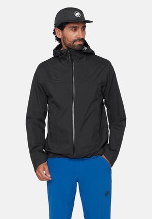 Mann trägt schwarze Kapuzenjacke und schwarze Kappe mit Tier-Logo, blaue Hose, steht mit Händen in den Taschen und schaut zur Seite.