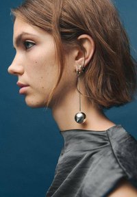 Larga arete plateado con un acento metálico esférico que cuelga de un alambre curvado; combinado con una prenda gris texturizada. Fondo azul.