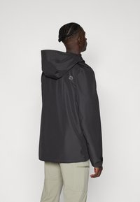 Veste imperméable gris foncé avec capuche, présentant un logo discret sur l'épaule et une texture lisse et brillante.