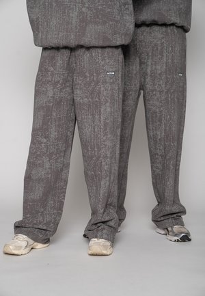 Two people wearing loose gray textured sweatpants and white sneakers standing side by side against a plain background.
