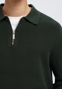 Hombre vistiendo polo de punto verde oscuro con cremallera plateada sobre camiseta interior blanca de cuello redondo contra fondo liso.