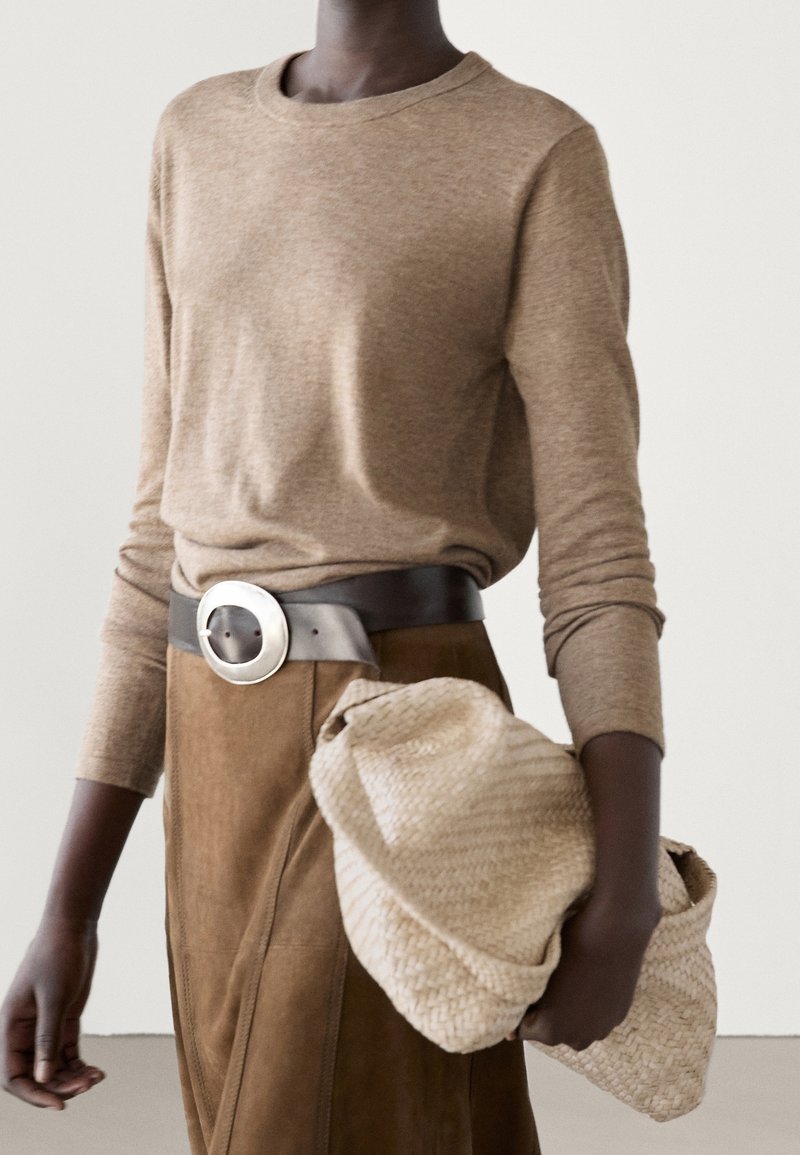Personne portant un haut beige à manches longues rentré dans une jupe en daim marron avec une ceinture noire à grande boucle argentée, tenant une pochette beige texturée.