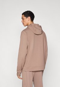 Καφέ φούτερ με κουκούλα, με ριμπ μανσέτες, άνετη εφαρμογή και ελαφρώς υφασμένο ύφασμα, εμφανιζόμενο από πίσω.