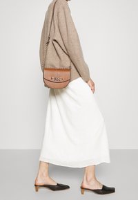 Borsa a tracolla in pelle marrone con tracolla a catena e logo impresso, forma arrotondata, finitura testurizzata, indossata con gonna crema e mules nere.