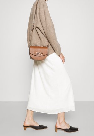 Braune Leder-Crossbody-Tasche mit Kettenriemen und geprägtem Logo, runde Form, strukturierte Oberfläche, getragen zu einem cremefarbenen Rock und schwarzen Mules.
