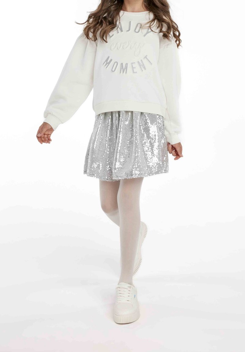 Fille marchant portant un sweat-shirt blanc avec l'inscription « Profite de chaque instant », une jupe en sequins argentés, des collants blancs et des baskets blanches sur fond blanc.