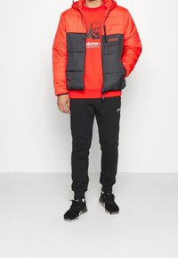 Vadderad jacka i stark röd med en svart mellandel, med en dragkedjeficka, bärs över en röd sweatshirt och svarta sweatpants med logotyper.