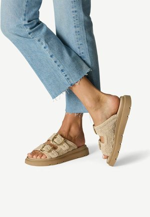 Sandales beige tissées style mule avec deux boucles dorées et une semelle rembourrée marron clair, associées à un jean bleu clair à ourlet effiloché.