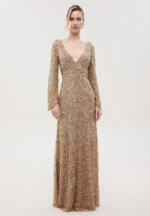 Femme portant une robe longue en sequins dorés à manches longues avec un travail de perles minutieux et un profond décolleté en V, debout devant un fond uni.