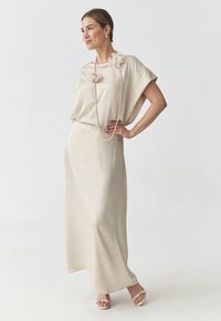 Robe beige clair avec des manches courtes et des embellissements floraux, présentant une silhouette droite et une texture satinée lisse. Une paire de sandales légères.