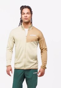 Man med flätat hår som bär en tvåtonad beige dragkedjetröja och mörkgröna Adidas Terrex-byxor, stående mot en vit bakgrund.