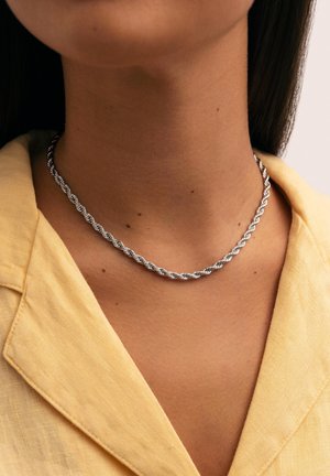 Collar de plata en forma de cuerda retorcida que se lleva alrededor del cuello, combinado con una camisa de color amarillo claro. Textura metálica suave, con un diseño simple.