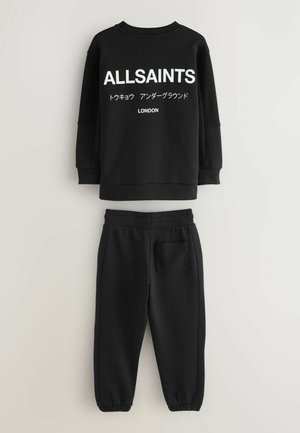 Melna sporta jaka ar tekstu "ALLSAINTS", japāņu rakstzīmēm un "LONDON" virsū, saskaņojamas melnas sporta bikses ar aizmugurējo kabatu.