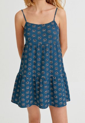 Robe sans manches bleue avec de fines bretelles et un motif floral blanc portée par une femme sur fond uni.