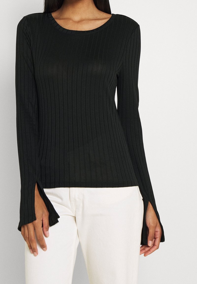 Černý žebrovaný top s dlouhým rukávem, který má kulatý výstřih, jemnou strukturu a rozparky na manžetách, v kombinaci se světle zbarvenými kalhotami.