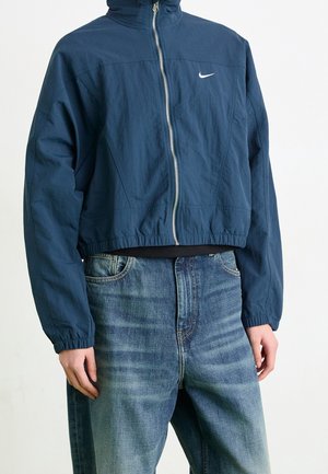 Persoon draagt een donkerblauw Nike-jas met ritssluiting en elastische manchetten, gecombineerd met lichtvervaagde blauwe jeans, staand tegen een effen achtergrond.