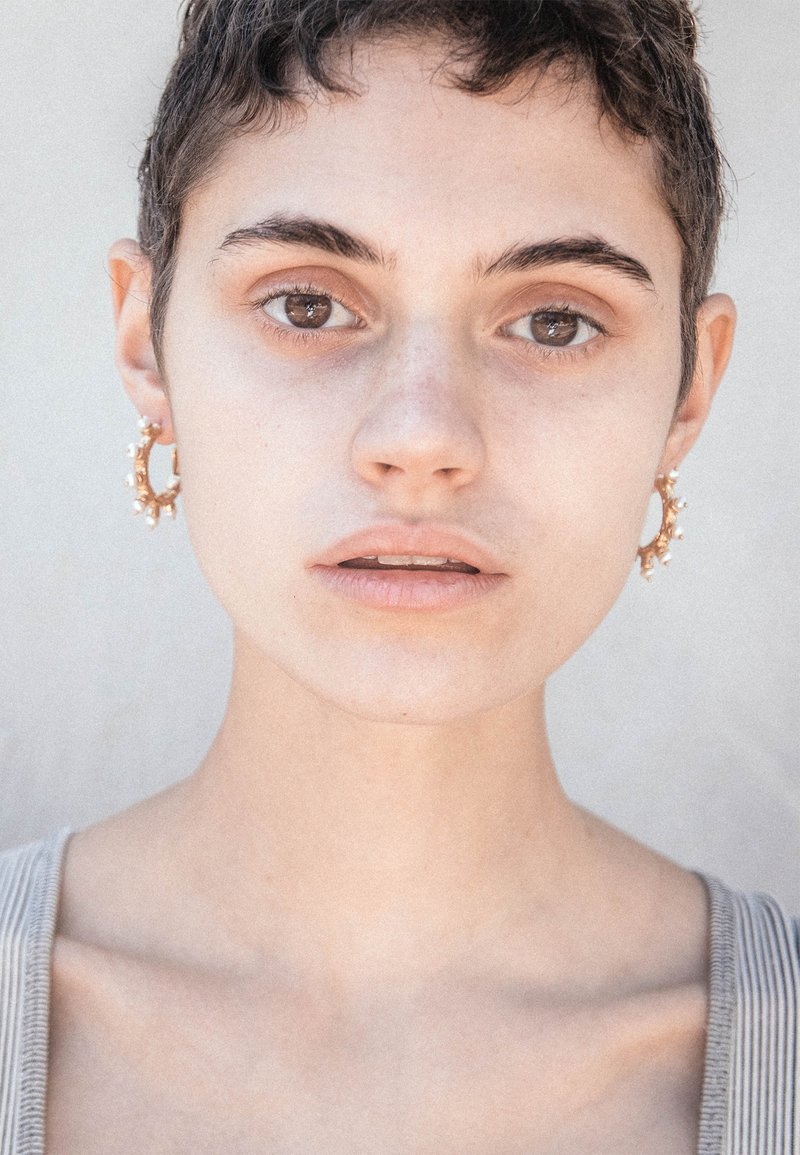 Aukso žiedinės auskarai su perlo akcentais, turintys tekstūruotą dizainą. Modelis turi trumpus plaukus ir neutralios spalvos topą be rankovių.