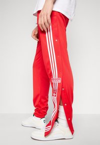 Osoba v červených teplákových kalhotách Adidas s bílými pruhy a částečně rozepnutými bočními knoflíky, doplněná o bílé tenisky a ponožky.