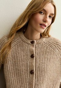 Cardigan tricoté en beige clair avec un col rond, comportant trois boutons marron foncé, un motif de point texturé et des manches raglan.