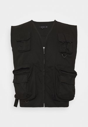 Gilet utilitaire noir sans manches avec plusieurs poches à l'avant, fermeture à glissière et anneaux métalliques sur un fond uni.