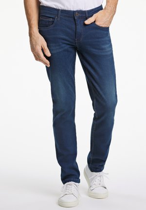 Vaqueros de mezclilla azul oscuro con un corte ajustado, sutil desgaste tipo whiskering y diseño clásico de cinco bolsillos, combinados con zapatillas blancas.